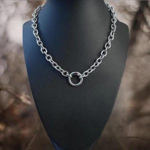 May include: A silver-colored chain necklace with a large circular pendant. The necklace is displayed on a black mannequin. The chain links are oval-shaped and evenly spaced. The background is blurred with neutral tones.