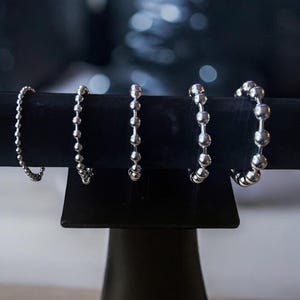 May include: Four silver ball chain bracelets of varying lengths displayed on a black stand. The bracelets are made of small, round metal beads.