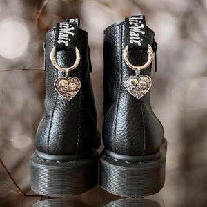 Puede incluir: Botas de cuero negro con un dije en forma de corazón con un diseño de calavera y huesos cruzados. El dije está unido a un anillo plateado con el texto "Little Miss" en él.
