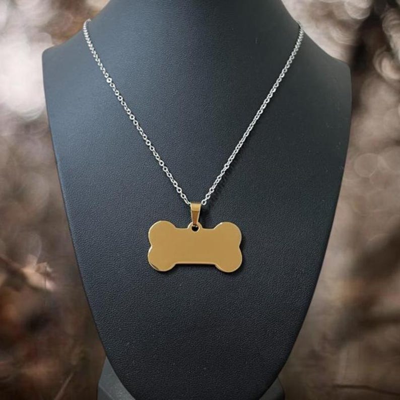 May include: A gold-colored, bone-shaped pendant hangs from a thin, silver-toned metal chain. The pendant has a smooth, polished surface with no visible engravings or markings. The chain features small, uniform links that connect to the top of the pendant.