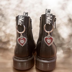 May include: Pair of black leather ankle boots with silver-tone hardware and a red heart charm with an eye design. The boots have a chunky sole and a pull tab with white text.