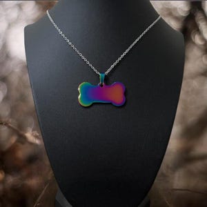 May include: A silver-toned chain necklace featuring a bone-shaped pendant with an iridescent, rainbow-colored finish that transitions from blue and green to purple and orange hues, suspended from a simple metal link chain.