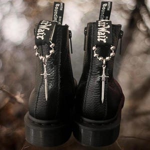 May include: Black leather ankle boots with a silver sword charm attached to a beaded chain. The boots have a zipper on the side and the text "With Sole Bouncing" on the back.