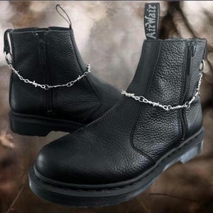 May include: A pair of black leather ankle boots with silver-colored barbed wire chain detailing. The boots have side zippers and pull tabs. The soles are thick and black, and the AirWair logo is visible on the back of one boot.