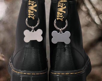Könnte beinhalten: Ein Paar schwarze Lederstiefel mit gelben Nähten und einem knochenförmigen Anhänger, der hinten hängt. Der Anhänger ist an einem silbernen Ring befestigt. Auf den Stiefeln steht in goldener Schrift "AirWair".