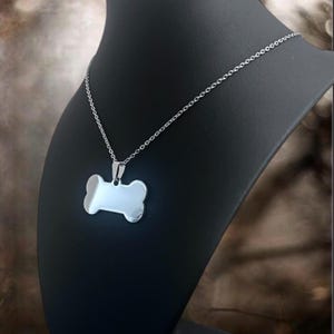 May include: A silver-toned metal necklace featuring a polished, smooth dog bone-shaped pendant suspended from a delicate link chain. The pendant has a reflective surface and is attached to the chain by a small, simple metal bail connector.