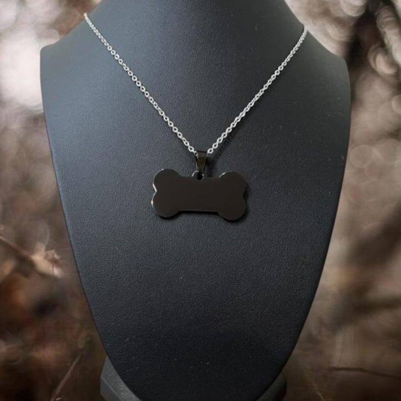 May include: A silver-toned metal chain necklace featuring a solid black, bone-shaped pendant. The pendant is suspended from a small metal bail attached to the center of the chain, creating a simple and clean design for this piece of jewelry.