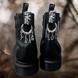 May include: Black leather ankle boots with silver charms spelling "YUNG BLUD". The boots feature a side zip and "Air Wair" text on the pull tab. The soles are black, and the boots have a textured finish. The boots are a stylish footwear choice.