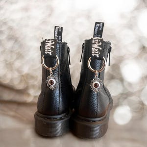 May include: Black leather ankle boots with a zipper on the side and a silver charm with an eye design hanging from the laces. The boots have a thick black sole and the laces have the text "With Soul Bouncing" on them.