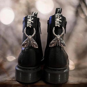 May include: Black leather ankle boots with a side zip and a silver moth charm attached to the zipper pull. The boots have a chunky black sole and the text "With Boudoir Wait" is printed on the zipper pull.