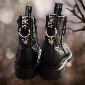 Puede incluir: Par de botines de cuero negro con detalles de cremallera plateada y un dije plateado adjunto a cada bota. El dije presenta una pequeña cabeza de gato detallada. Las botas tienen una etiqueta negra y blanca que dice "Air Wait" con el texto "With Sole & Bouncing" arriba.