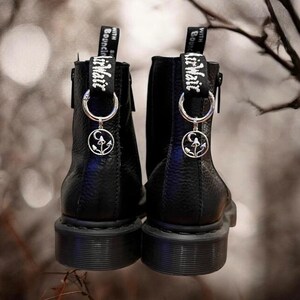 May include: A pair of black leather ankle boots with silver charms attached to the laces. The charms feature a crescent moon and mushrooms. The boots have a zipper on the inside of each ankle and a black strap with the text "Lit Wait" and "With a Bounce" on each strap.