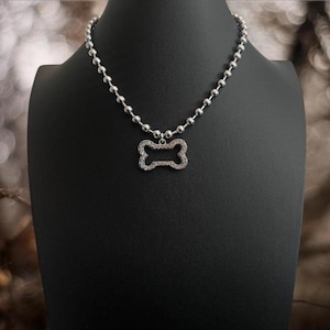 May include: A silver-colored dog bone pendant necklace. The pendant is encrusted with small, clear stones and hangs from a beaded chain. The necklace is displayed on a black mannequin.