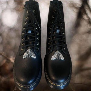 May include: Pair of black leather combat boots with silver moth embellishments on the toes. The boots have black laces, gold eyelets, and a thick, black sole. A stylish and edgy footwear choice.