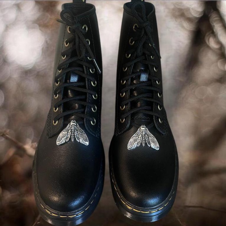 May include: Pair of black leather combat boots with silver moth embellishments on the toes. The boots have black laces, gold eyelets, and a thick, black sole. A stylish and edgy footwear choice.