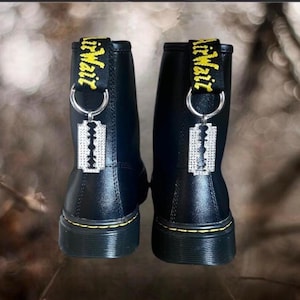 May include: Pair of black leather ankle boots with yellow stitching and a yellow and black pull tab that says "Air Wair". Each boot has a silver charm in the shape of a razor blade.