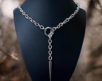 May include: A silver-colored necklace with a chain design. The necklace features a large ring in the center, with a chain that hangs down and ends in a pointed spike. The necklace is displayed on a black mannequin.