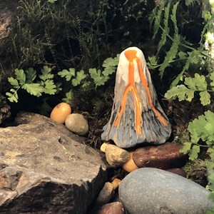 May include: A miniature ceramic volcano with orange lava flowing down the sides. The volcano is surrounded by small rocks and green foliage.