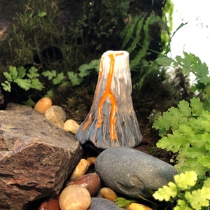 May include: A small ceramic volcano with orange lava flowing down the sides, surrounded by rocks and greenery.