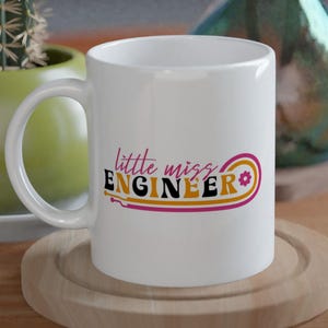 May include: White ceramic mug with the text "little miss ENGINEER" in pink, orange, and yellow. The mug is on a wooden coaster on a wooden table. A cactus in a green pot and a decorative vase are in the background.