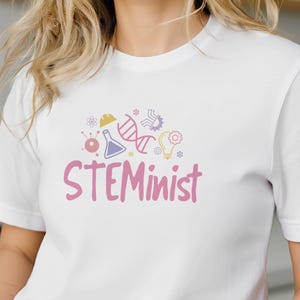 May include: White t-shirt featuring the word "STEMinist" in pink, accompanied by a colourful graphic of science symbols. The shirt is made of a soft material, suitable for casual wear.