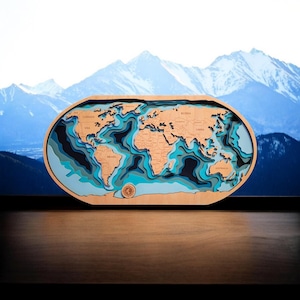 May include: A wooden, layered world map with a light brown frame. The map features detailed coastlines and country names, with varying shades of blue representing ocean depths. The background shows a mountain range.