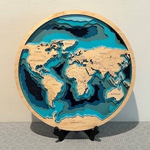 May include: A wooden world map with layered continents and oceans in shades of blue. The map is mounted on a black stand and features labels for countries like Canada, United States, Brazil, Russia, China, and Australia. The map also includes the text "ANTARCTICA" at the bottom.