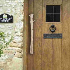 Peut inclure: Une porte en bois à panneaux verticaux, une petite fenêtre carrée à quatre carreaux, un heurtoir en métal noir, une fente à courrier noire et une longue poignée en bois clair. Une plaque noire à gauche indique THE CORNER HOUSE.