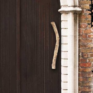 Peut inclure: Une double porte en bois cintrée de couleur brun foncé, insérée dans un cadre en pierre claire. La porte présente des panneaux verticaux et une poignée en bois clair et incurvée fixée sur le côté droit. Elle est entourée d'un mur en briques rouges.