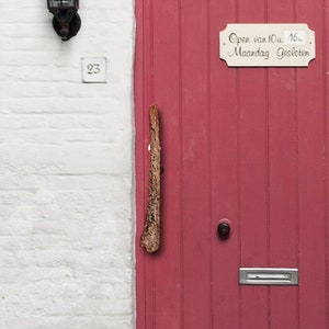 Könnte beinhalten: Eine rote Holztür mit einem Schild mit der Aufschrift "Open van 10u. 16u. Maandag Gesloten". Die Tür hat einen Holzgriff und einen silbernen Briefkasten.