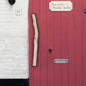 Può includere: Una porta verticale in legno rosso con una maniglia a ramo naturale chiaro, una fessura per la posta argentata, un pomello rotondo e un cartello rettangolare con il testo Open van 10u. 16. Maandag Gesloten. Due lanterne nere fiancheggiano la porta.