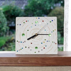 May include: Square white clock with a decorative face featuring concentric circles and colourful embellishments. The clock has black hands and a gold-tone centre, with green, blue, and red accents. The clock is sitting on a wooden surface.
