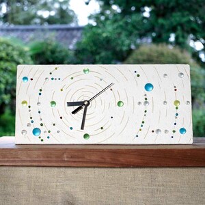 May include: A rectangular white clock with a spiral design and colorful glass beads. The clock hands are black. The clock is sitting on a wooden surface, with a green background.