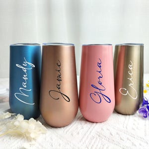 May include: Four stemless champagne flutes in teal, copper, pink, and gold. Each tumbler has a name in cursive script: Mandy, Jamie, Gloria, and Erica. The flutes are on a white surface with floral accents.