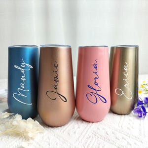 May include: Four stemless champagne flutes in teal, copper, pink, and gold. Each flute has a name in cursive script: Mandy, Jamie, Gloria, and Erica. The flutes are on a white surface with floral accents.