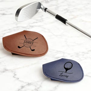 Puede incluir: Dos fundas personalizadas para la cabeza del palo de golf, una marrón y otra azul marino, con un diseño de palo y bola de golf. La funda marrón tiene el nombre "James" y la funda azul marino tiene el nombre "Lucas". Un palo de golf está en el fondo.