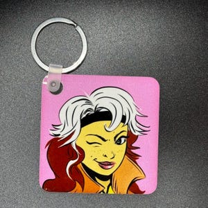 May include: A pink keychain with a cartoon image of a woman with white hair, a yellow headband, and a wink. The woman is wearing a brown and orange jacket.