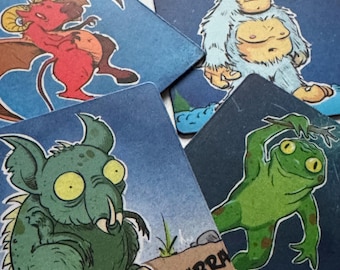 Könnte beinhalten: Vier blaue quadratische Untersetzer mit Illustrationen von Fabelwesen. Die Untersetzer zeigen einen roten Drachen, einen weißen Yeti, einen grünen Frosch und einen grünen Chupacabra.