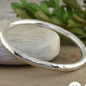 Puede incluir: Un brazalete de plata martillada. El brazalete tiene una superficie texturizada y una forma redondeada. El fondo incluye una piedra de color claro y follaje verde, sobre una superficie de madera.