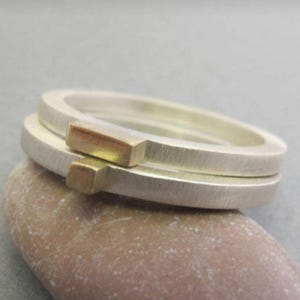 Könnte beinhalten: Zwei silberne Ringe mit gebürsteter Oberfläche, jeweils mit einem kleinen, rechteckigen Goldakzent. Die Ringe sind übereinander gestapelt und zeigen ein minimalistisches Design. Die Ringe liegen auf einem hellbraunen Stein. Maße in Zentimetern.