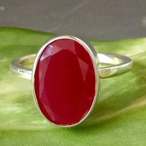 Könnte beinhalten: Ein silberner Ring mit einem großen, oval geformten, tiefroten Edelstein. Der Edelstein ist facettiert und fängt das Licht ein. Der Ring hat ein schlichtes, poliertes Silberband. Steinabmessungen ca. 2 cm x 1,5 cm.