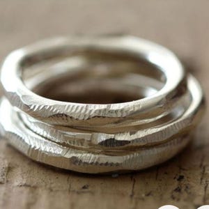May include: Close-up of three silver rings stacked together. Each ring has a textured, hammered surface, creating a unique, organic look. The rings are made of a shiny, metallic material, reflecting light. The rings are a piece of jewelry.