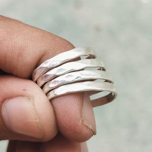 Puede incluir: Un conjunto de cinco anillos de plata martillada. Cada anillo presenta una superficie texturizada e irregular, creando una apariencia única y artesanal. Los anillos se llevan en un dedo, mostrando su diseño simple pero elegante. La plata refleja la luz.
