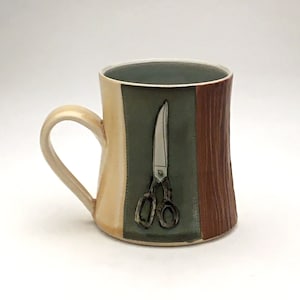 Könnte beinhalten: Eine Keramiktasse mit einem beige, grünen und braunen Design. Die Tasse zeigt eine Schere in der Mitte des Designs.