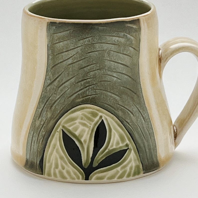 K&ouml;nnte beinhalten: Eine handgefertigte Keramik-Tasse mit einem einzigartigen Design. Die Tasse hat eine beige Au&szlig;enseite mit einem zentralen, strukturierten gr&uuml;nen Feld und einem Blattmotiv. Der Griff und der Rand sind ebenfalls beige, was einen k&uuml;nstlerischen Look erzeugt.