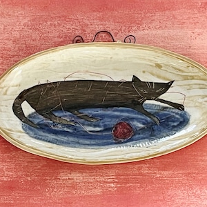 Könnte beinhalten: Eine ovale Servierplatte aus Keramik mit einem schwarzen Katzenmotiv. Die Katze liegt und spielt mit einem roten Wollknäuel. Die Platte hat einen blauen und weißen Hintergrund.
