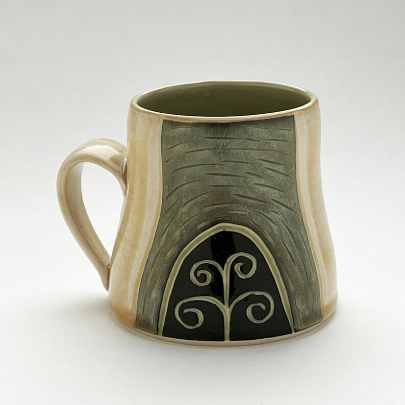 K&ouml;nnte beinhalten: Handgefertigte Keramiktasse mit beigefarbenem &Auml;u&szlig;eren und gr&uuml;nem Inneren. Die Tasse hat ein strukturiertes, dunkelgr&uuml;nes Design auf der Vorderseite mit einem schwarzen, stilisierten Blumenmotiv. Der Henkel ist beige, und die Tasse hat eine abgerundete Form, ca. 10 cm hoch.