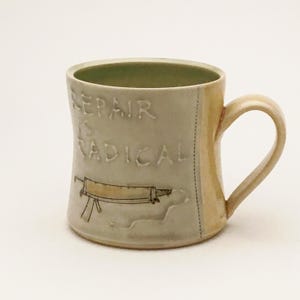 Könnte beinhalten: Eine Keramiktasse mit hellbeigem Glase und gelbem Glase am Henkel. Die Tasse trägt den Text "Repair is Radical" in braunem Glase auf der Vorderseite. Unter dem Text befindet sich eine stilisierte Zeichnung eines Werkzeugs.