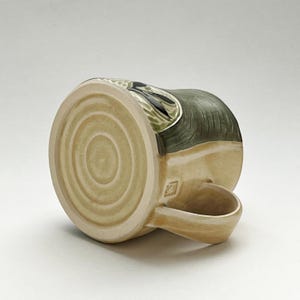 K&ouml;nnte beinhalten: Eine handgefertigte Keramiktasse mit beigem Boden und dunkelgr&uuml;nem Oberteil. Die Tasse hat einen gebogenen Henkel und ein dekoratives Design an der Seite. Der Boden der Tasse hat konzentrische Kreise.