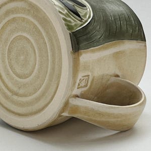 K&ouml;nnte beinhalten: Eine Keramik-Tasse mit beigefarbenem &Auml;u&szlig;eren und einem dunkelgr&uuml;nen und schwarzen Design. Die Tasse hat einen abgerundeten Henkel und einen kreisf&ouml;rmigen Boden mit konzentrischen Ringen. Ein kleines quadratisches Emblem ist in der N&auml;he des Henkels zu sehen.